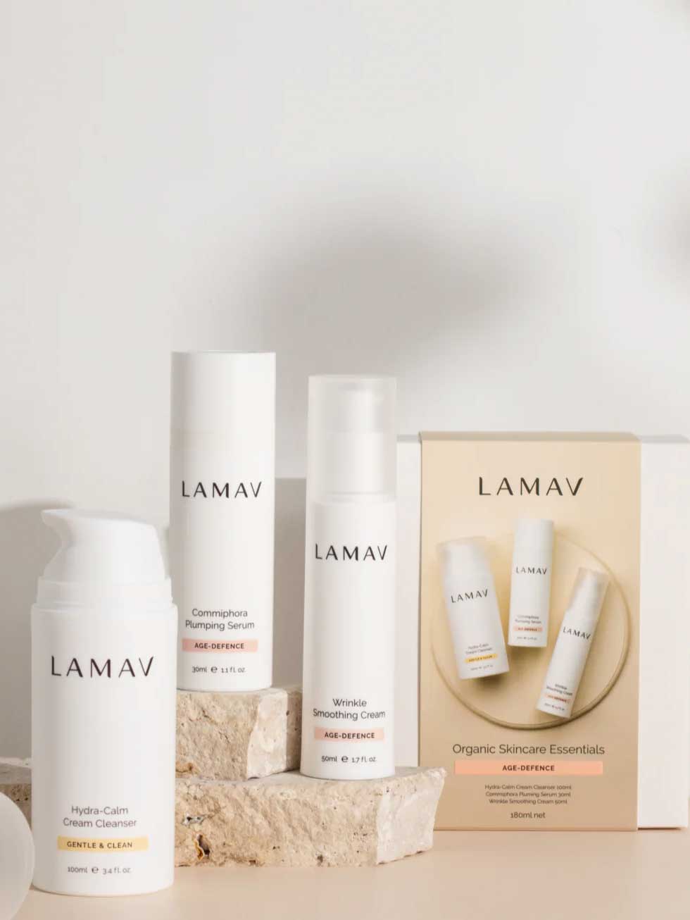 Three LAMAV Age-Defence Skin Discovery Pack products are displayed on stone blocks, with a branded box labeled "Organic Skincare Essentials Age-Defence" showcasing the anti-ageing range against a neutral background.