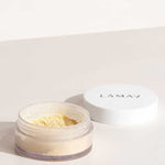 An open LAMAV Mattifying Powder 3g jar with a white lid rests on a light surface. The clear container reveals some of the yellow-tinted, talc-free powder inside.