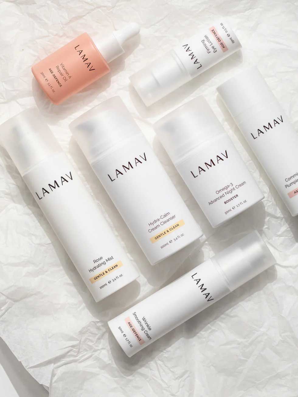 Several white and pink bottles from the LAMAV Age-Defence Complete Skincare Collection for Dry/Sensitive Skin (7 pack) are arranged on crumpled white paper—organic creams, mist, and serums ideal for sensitive skin.
