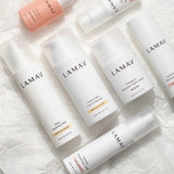 Several white and pink bottles from the LAMAV Age-Defence Complete Skincare Collection for Dry/Sensitive Skin (7 pack) are arranged on crumpled white paper—organic creams, mist, and serums ideal for sensitive skin.
