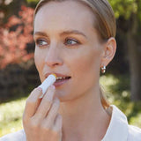 A woman in a white shirt and silver hoop earrings applies Lavera Basis Sensitiv Lip Balm 4.5g outdoors. With light hair and blue eyes, she enjoys the sunlight; the balm's organic jojoba oil gently nourishes her sensitive lips.
