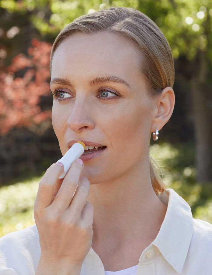 A woman in a white shirt and silver hoop earrings applies Lavera Basis Sensitiv Lip Balm 4.5g outdoors. With light hair and blue eyes, she enjoys the sunlight; the balm's organic jojoba oil gently nourishes her sensitive lips.