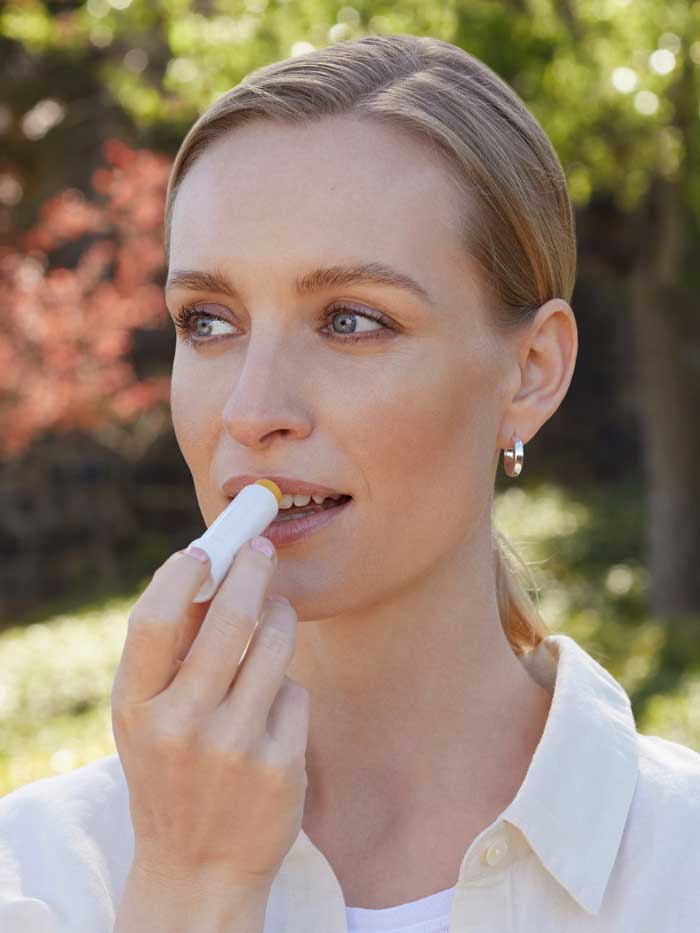 A woman in a white shirt and silver hoop earrings applies Lavera Basis Sensitiv Lip Balm 4.5g outdoors. With light hair and blue eyes, she enjoys the sunlight; the balm's organic jojoba oil gently nourishes her sensitive lips.