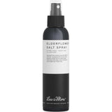 A black 150ml spray bottle labeled "Less is More Elderflower Salt Spray" features a white label. This texturising spray is ideal for hair styling and creating effortless beach waves.