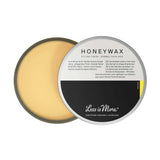 A round, open container of Less is More Honeywax 50g reveals yellowish organic wax inside. The partially covered lid features black and grey text detailing its natural hold formula.