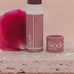 Uncapped Biode Lilly Pilly balm with a deep plum-coloured stick and rose-pink paper tube on pink stone, beside its cap and a rose petal. Lilly Pilly is printed below.