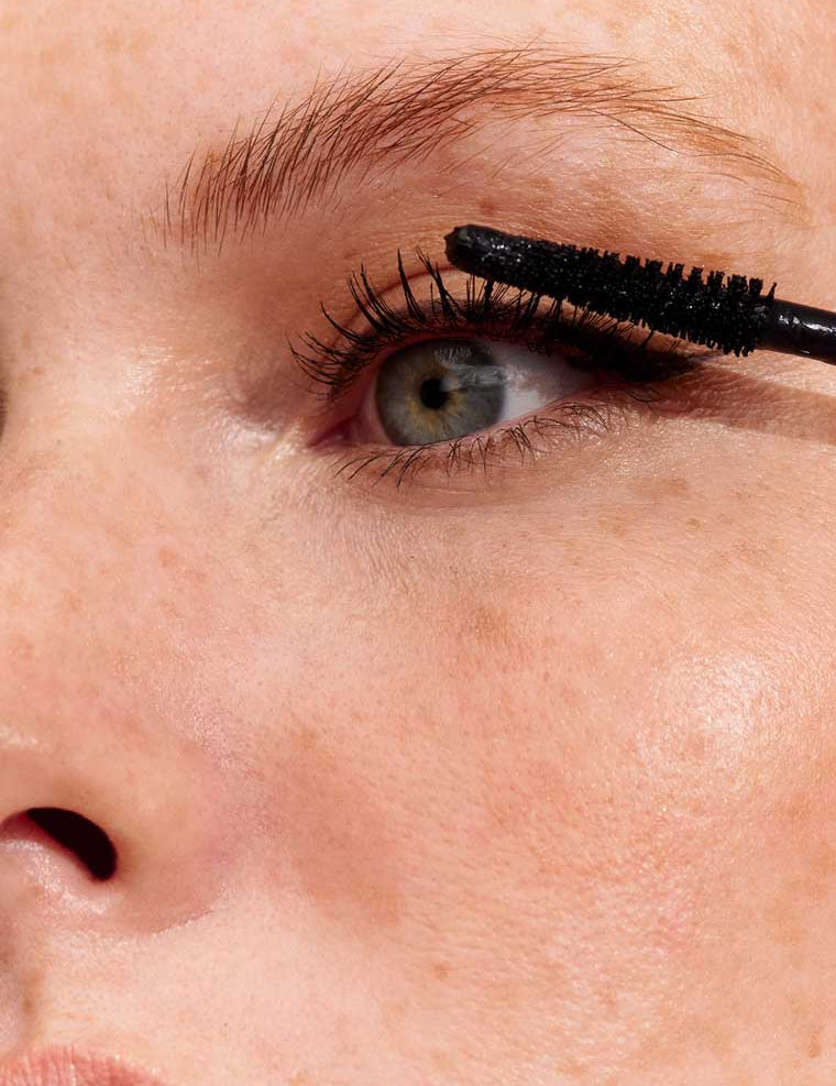 Close-up of a person applying INIKA Organic Long Lash Mascara 8ml to their upper lashes. The image highlights the eye, eyebrow, part of the cheek, clear skin, and visible freckles.