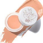 An open pot of Golden Peach Long Last Concealer rests on a matching golden peach smear. The white lid beside the pot has a fine botanical drawing of leaves and berries.