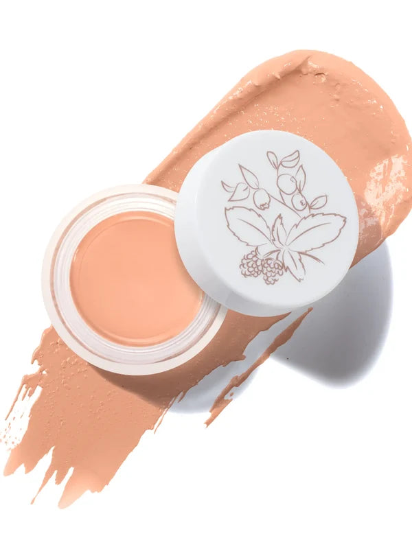 An open pot of Golden Peach Long Last Concealer rests on a matching golden peach smear. The white lid beside the pot has a fine botanical drawing of leaves and berries.