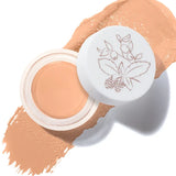 An open pot of Peach Bisque Long Last Concealer rests on a matching warm peach beige smear. The white lid beside the pot has a fine botanical drawing of leaves and berries.