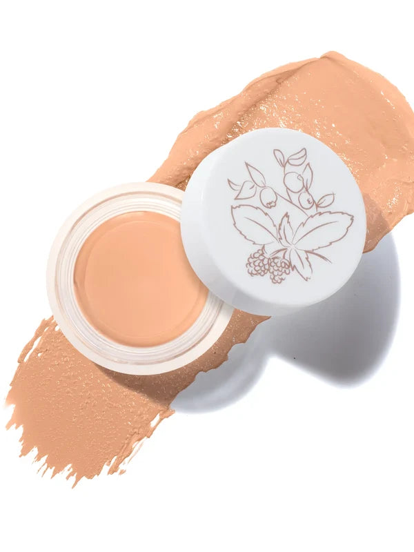 An open pot of Peach Bisque Long Last Concealer rests on a matching warm peach beige smear. The white lid beside the pot has a fine botanical drawing of leaves and berries.