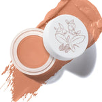 An open pot of Toffee Long Last Concealer rests on a matching warm tan smear. The white lid beside the pot has a fine botanical drawing of leaves and berries.