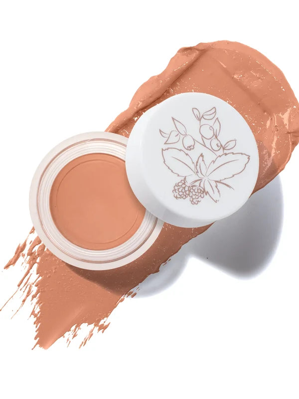 An open pot of Toffee Long Last Concealer rests on a matching warm tan smear. The white lid beside the pot has a fine botanical drawing of leaves and berries.