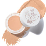 An open pot of White Peach Long Last Concealer rests on a matching peach beige smear. The white lid beside the pot has a fine botanical drawing of leaves and berries.