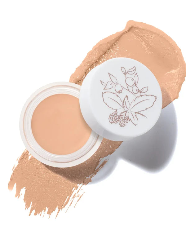 An open pot of White Peach Long Last Concealer rests on a matching peach beige smear. The white lid beside the pot has a fine botanical drawing of leaves and berries.