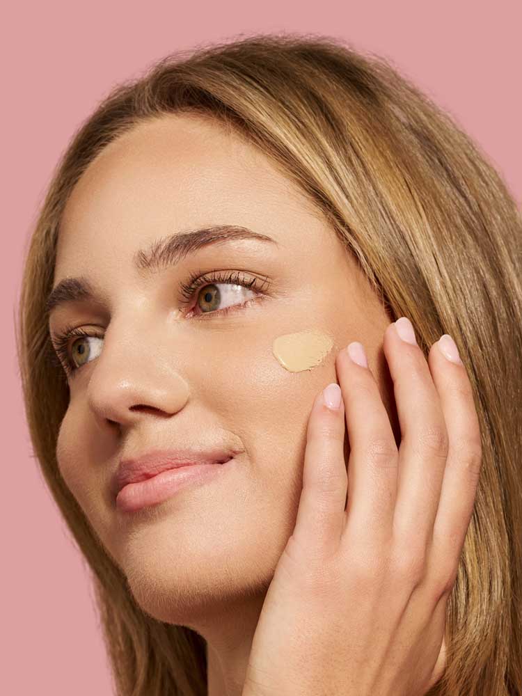 A woman with light brown hair applies The Jojoba Company Illuminating Tinted Moisturiser SPF15 50ml to her cheek with her fingers, smiling softly against a solid pink background.