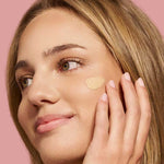 A woman with light brown hair applies The Jojoba Company Illuminating Tinted Moisturiser SPF15 50ml to her cheek with her fingers, smiling softly against a solid pink background.