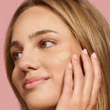 A woman with light brown hair applies The Jojoba Company Illuminating Tinted Moisturiser SPF15 50ml to her cheek with her fingers, smiling softly against a solid pink background.