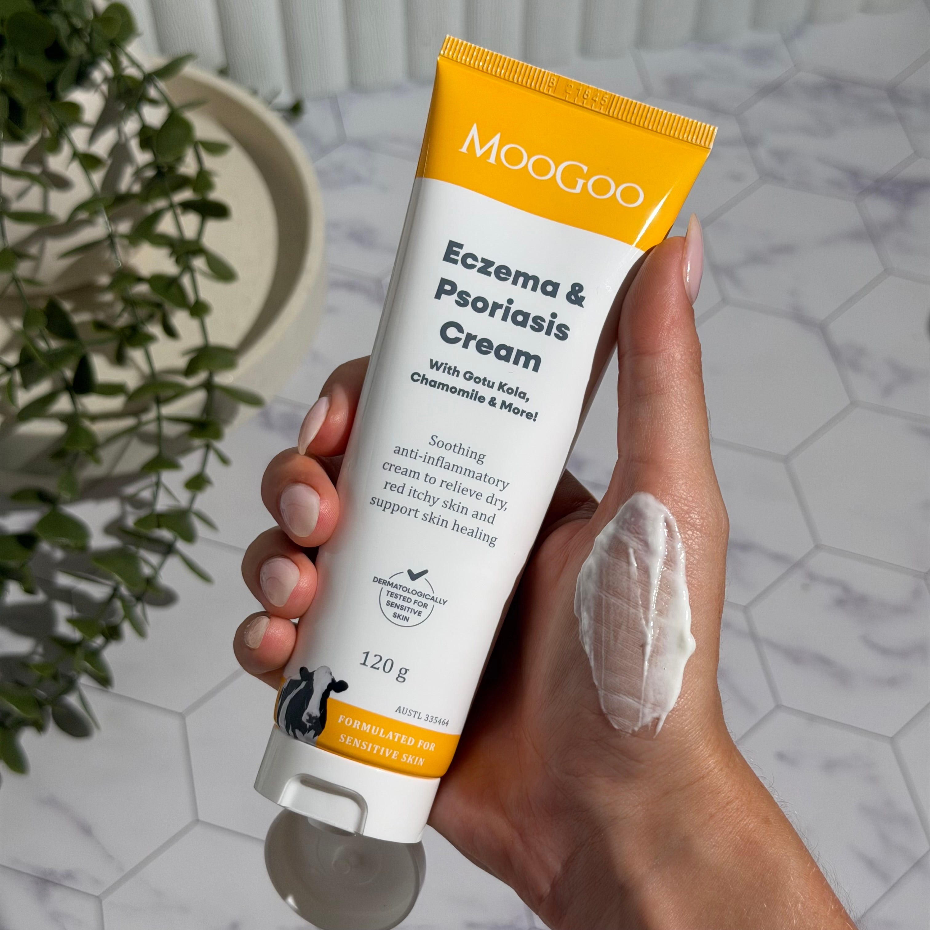 Hand holding an open white-and-yellow MooGoo Eczema and Psoriasis Cream tube marked 120g, with a thick white smear on the back of the hand.