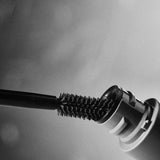 A close-up, black-and-white shot shows the Madara Bold Volume Mascara - Deep Matter 6ml wand being pulled from its tube, bristles coated. A soft-focus background enhances the mascara’s pure, elegant appeal.