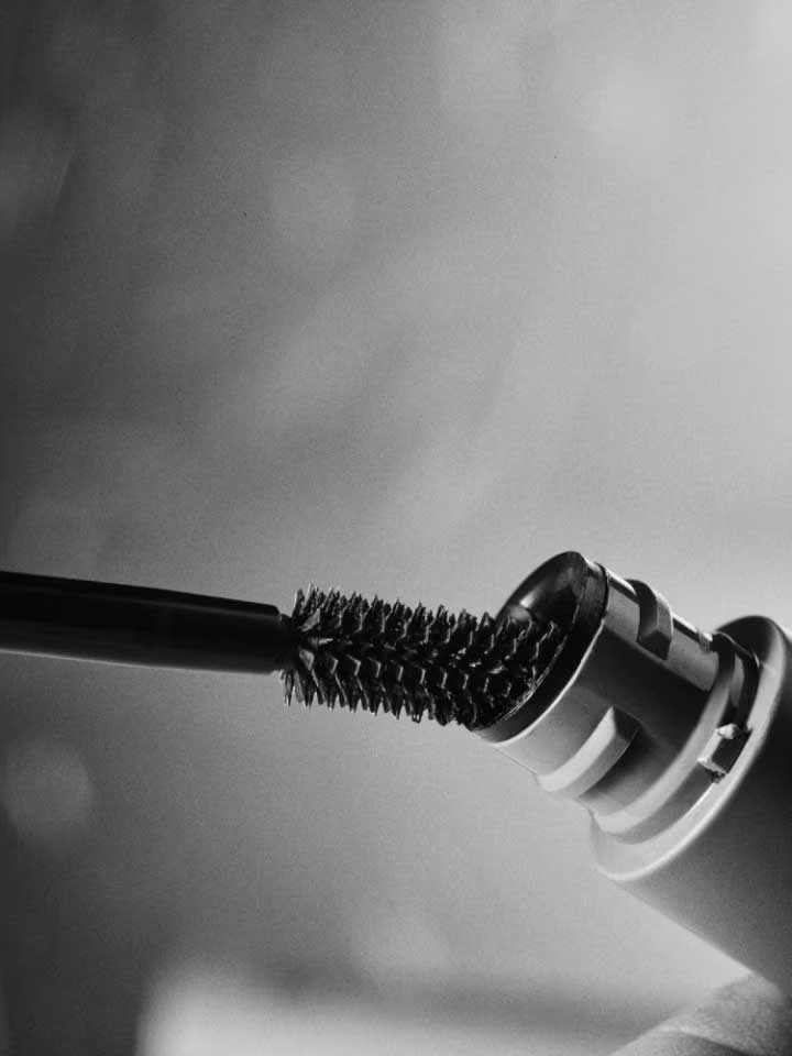 A close-up, black-and-white shot shows the Madara Bold Volume Mascara - Deep Matter 6ml wand being pulled from its tube, bristles coated. A soft-focus background enhances the mascara’s pure, elegant appeal.