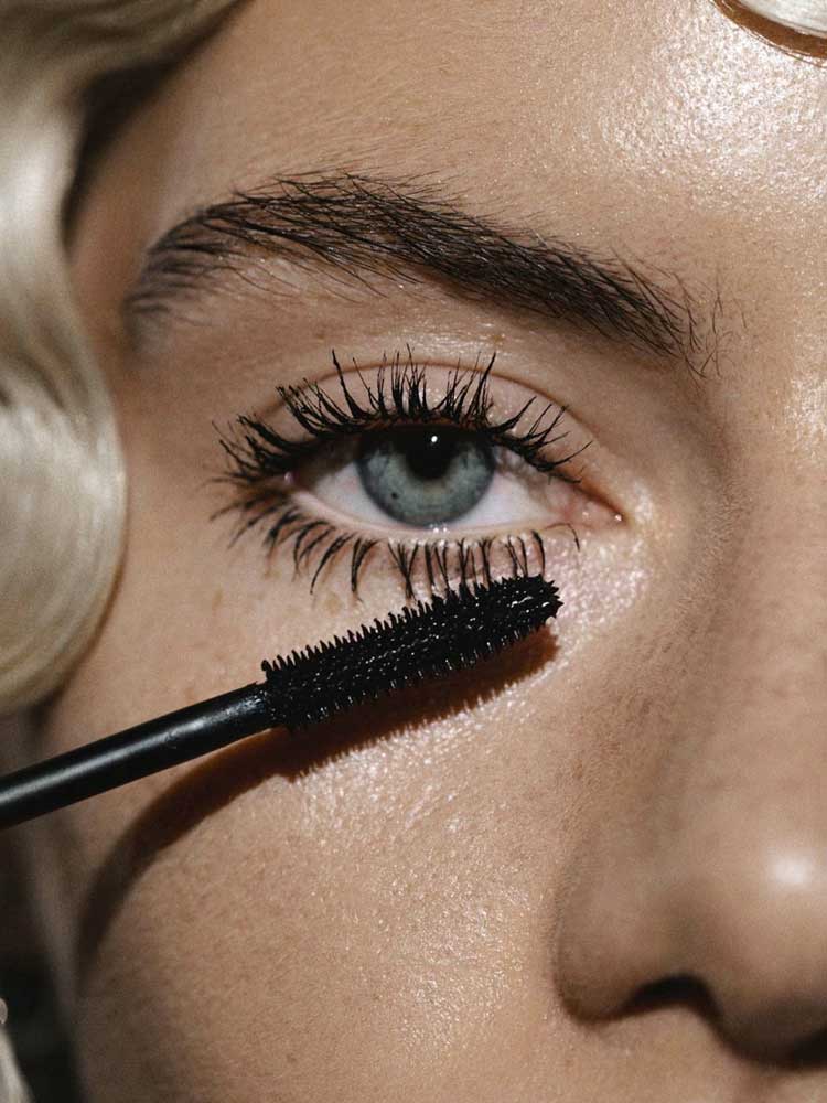 Close-up of a light-skinned person with blonde hair applying Madara Bold Volume Mascara - Deep Matter 6ml to long upper lashes on one blue eye. A vegan mascara wand coats the lashes in black.
