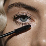 Close-up of a light-skinned person with blonde hair applying Madara Bold Volume Mascara - Deep Matter 6ml to long upper lashes on one blue eye. A vegan mascara wand coats the lashes in black.