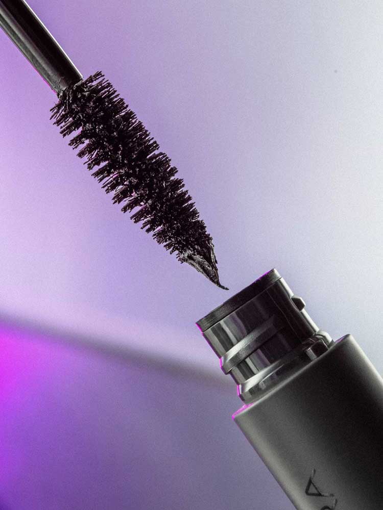 Close-up of the Madara Come Closer Infinite Lash Mascara - Black 6ml wand with black formula on the bristles, held above its open tube against a softly lit purple-toned background.