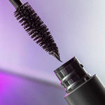Close-up of the Madara Come Closer Infinite Lash Mascara - Black 6ml wand with black formula on the bristles, held above its open tube against a softly lit purple-toned background.