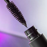 Close-up of the Madara Come Closer Infinite Lash Mascara - Black 6ml wand with black formula on the bristles, held above its open tube against a softly lit purple-toned background.