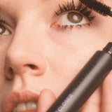 A person applies Madara Come Closer Infinite Lash Mascara - Black 6ml to their upper lashes. The tube, labeled "COME CLOSER" in purple, is held near their face, with attention on the eye and elongated lashes.