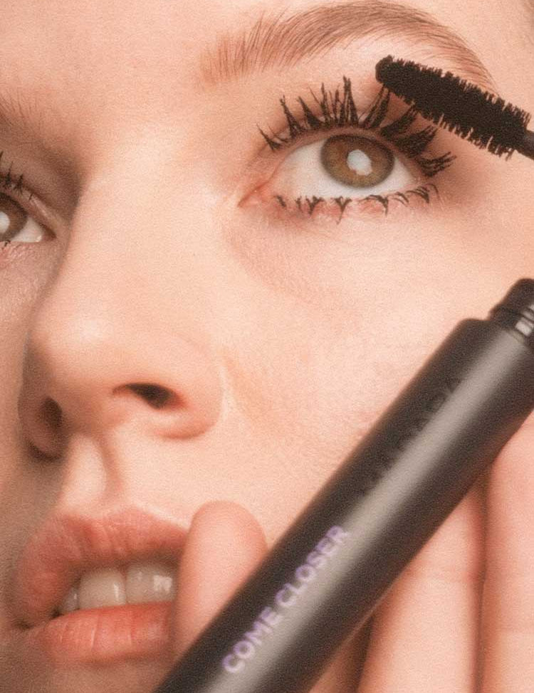A person applies Madara Come Closer Infinite Lash Mascara - Black 6ml to their upper lashes. The tube, labeled "COME CLOSER" in purple, is held near their face, with attention on the eye and elongated lashes.