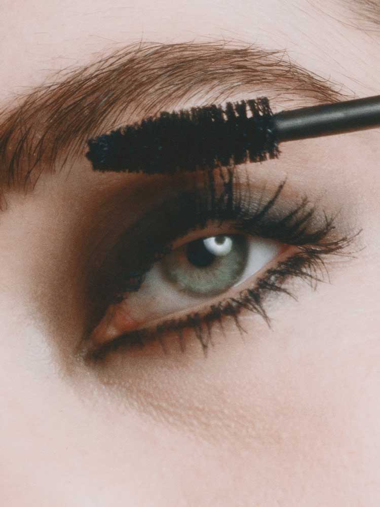 A close-up of a person’s eye with dramatic makeup, applying Madara Come Closer Infinite Lash Mascara - Black 6ml to their upper lashes using a mascara wand.