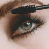 A close-up of a person’s eye with dramatic makeup, applying Madara Come Closer Infinite Lash Mascara - Black 6ml to their upper lashes using a mascara wand.