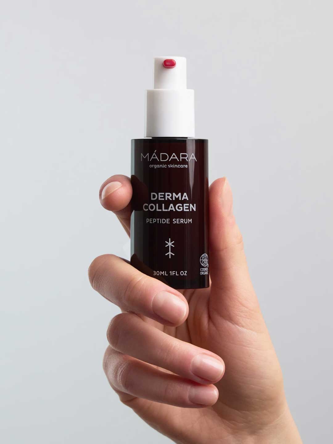 A hand holds a brown bottle of Madara Derma Collagen Peptide Serum 30ml with a white pump, dispensing some of this organic, anti-aging serum. The background is plain and light-colored.