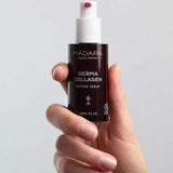 A hand holds a brown bottle of Madara Derma Collagen Peptide Serum 30ml with a white pump, dispensing some of this organic, anti-aging serum. The background is plain and light-colored.