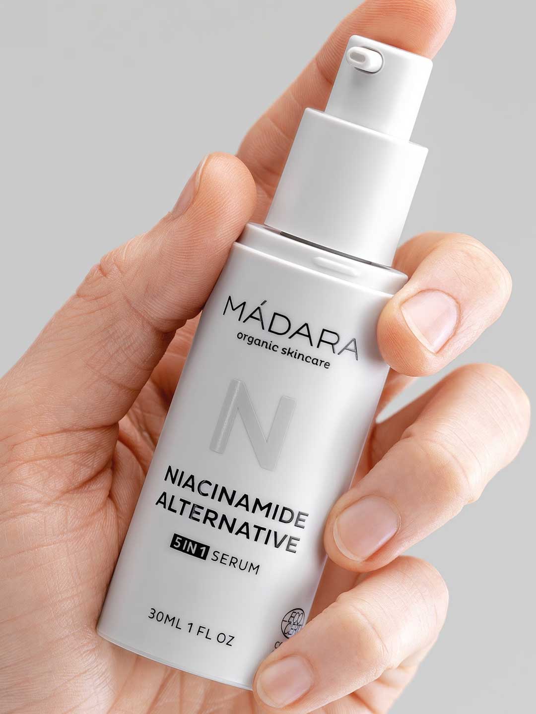 A hand holds a white bottle of Madara Niacinamide Alternative 5-in-1 Serum 30ml with a pump dispenser and minimalist design, set against a light gray background. Perfect for breakout-prone skin.