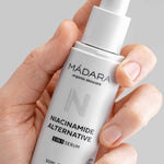 A hand holds a white bottle of Madara Niacinamide Alternative 5-in-1 Serum 30ml with a pump dispenser and minimalist design, set against a light gray background. Perfect for breakout-prone skin.