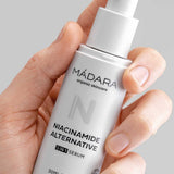 A hand holds a white bottle of Madara Niacinamide Alternative 5-in-1 Serum 30ml with a pump dispenser and minimalist design, set against a light gray background. Perfect for breakout-prone skin.