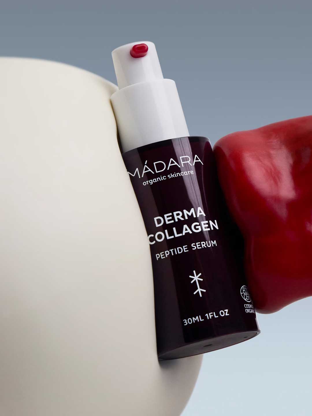 A 30ml bottle of Madara Derma Collagen Peptide Serum is held between a white surface and a shiny red object, set against a light blue background. The dark bottle features white text and a white pump.
