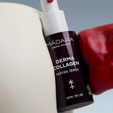 A 30ml bottle of Madara Derma Collagen Peptide Serum is held between a white surface and a shiny red object, set against a light blue background. The dark bottle features white text and a white pump.