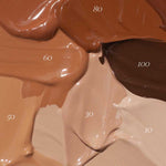 Thick swatches of Madara Skin Equal Soft Glow Foundation SPF15 30ml in shades 10 to 100 are arranged in a textured, overlapping display, highlighting the product’s diverse range of skin tone options.
