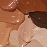 Thick swatches of Madara Skin Equal Soft Glow Foundation SPF15 30ml in shades 10 to 100 are arranged in a textured, overlapping display, highlighting the product’s diverse range of skin tone options.