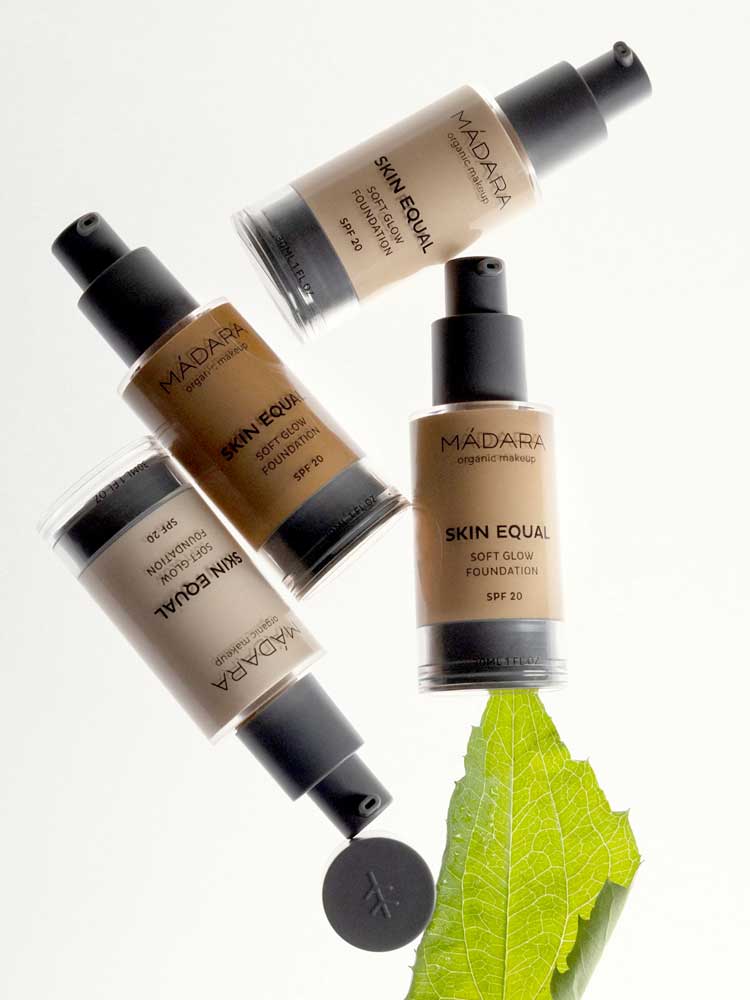 Four bottles of Madara Skin Equal Soft Glow Foundation SPF15 30ml are arranged on a white background, with one bottle resting on a green leaf.