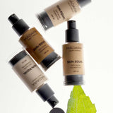 Four bottles of Madara Skin Equal Soft Glow Foundation SPF15 30ml are arranged on a white background, with one bottle resting on a green leaf.