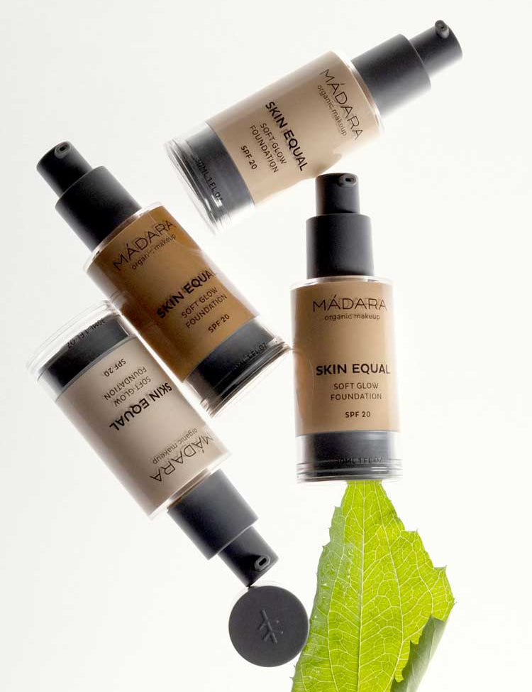 Four bottles of Madara Skin Equal Soft Glow Foundation SPF15 30ml are arranged on a white background, with one bottle resting on a green leaf.