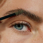 A close-up shows tinted brow gel being applied to a person’s eyebrow with a spoolie, grooming the hairs. The skin displays freckles and fine hairs. Product: Madara Grow & Fix Tinted Brow Gel 4.25ml.
