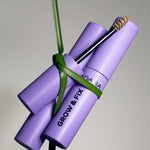 Three Madara Grow & Fix Tinted Brow Gel 4.25ml tubes in lavender are bundled with a green leaf. One tube is open, showing the spoolie brush with tinted brow gel on its bristles.