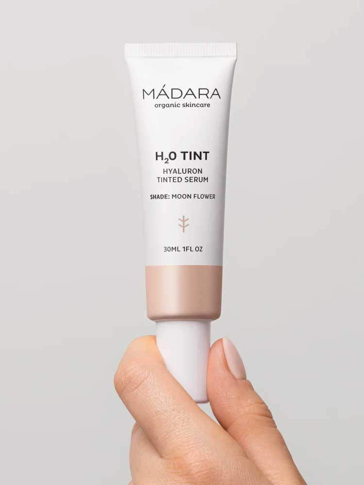 A hand holds Madara H2O Tint Hyaluron Tinted Serum 30ml in shade Moon Flower, a hyaluronic acid-infused tinted moisturiser, against a neutral background.