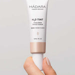 A hand holds Madara H2O Tint Hyaluron Tinted Serum 30ml in shade Moon Flower, a hyaluronic acid-infused tinted moisturiser, against a neutral background.